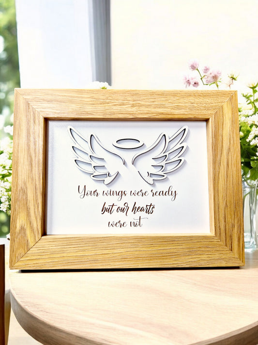 Memorial wooden frame with glitter angel wings and engraved verse, displayed on a table with flowers in the background.