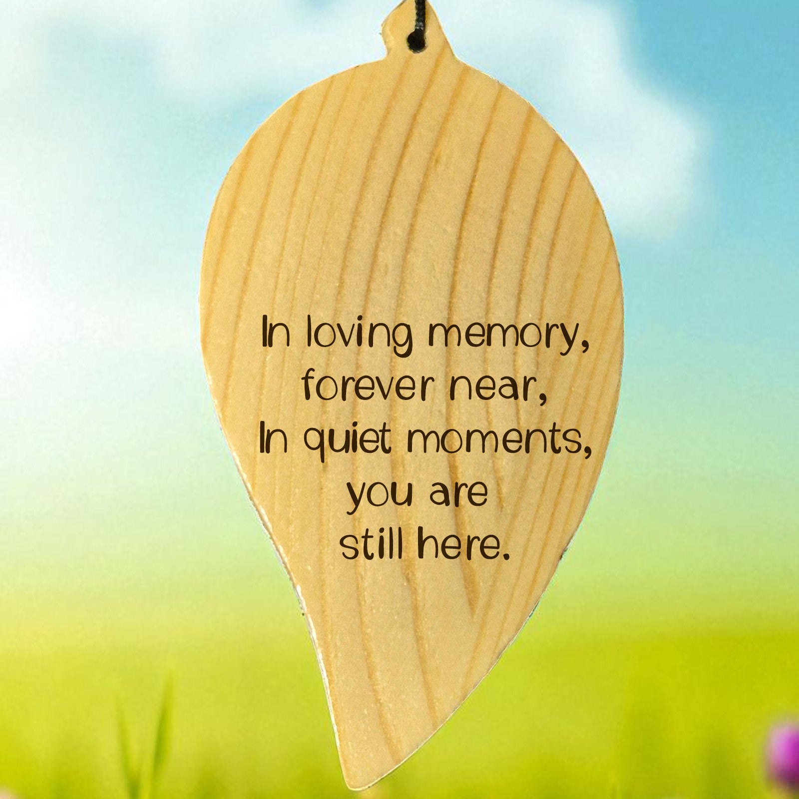 Wooden engraved leaf-shaped wind chime with memorial quote on it, set against a blurred natural background.