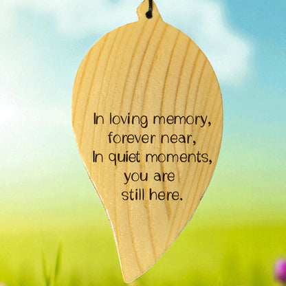Wooden engraved leaf-shaped wind chime with memorial quote on it, set against a blurred natural background.