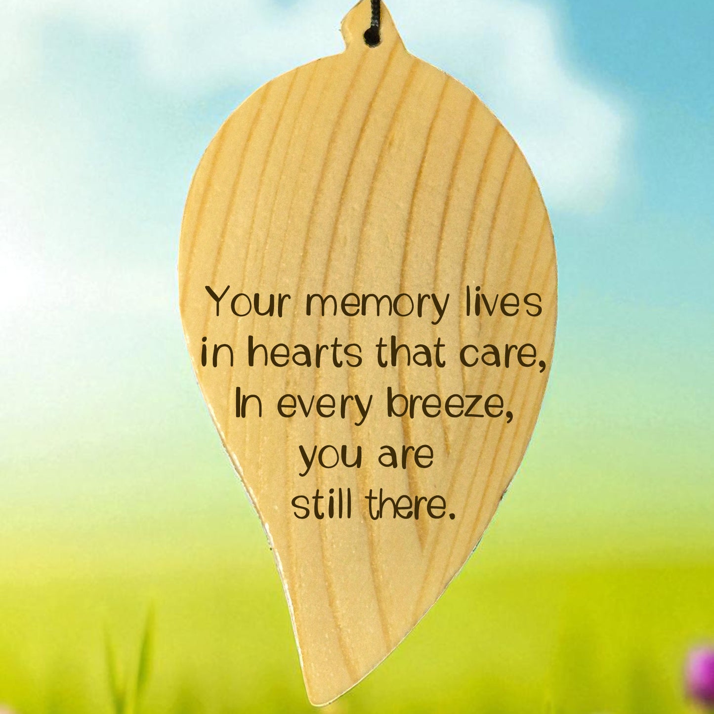 Wooden engraved leaf-shaped wind chime with a memorial quote on it, set against a blurred natural background.