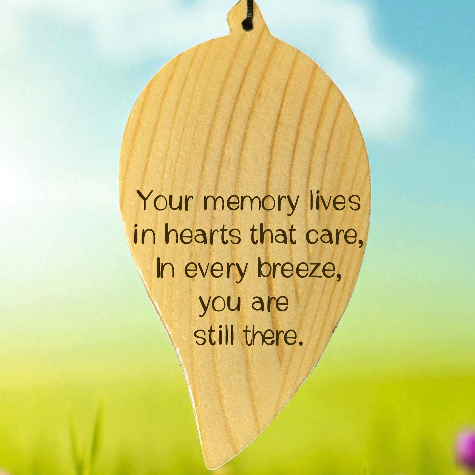 Wooden engraved leaf-shaped wind chime with a memorial quote on it, set against a blurred natural background.