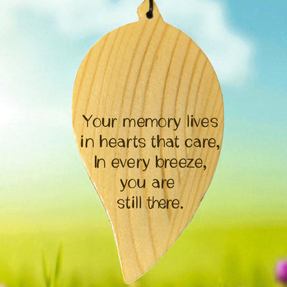 Wooden engraved leaf-shaped wind chime with a memorial quote on it, set against a blurred natural background.