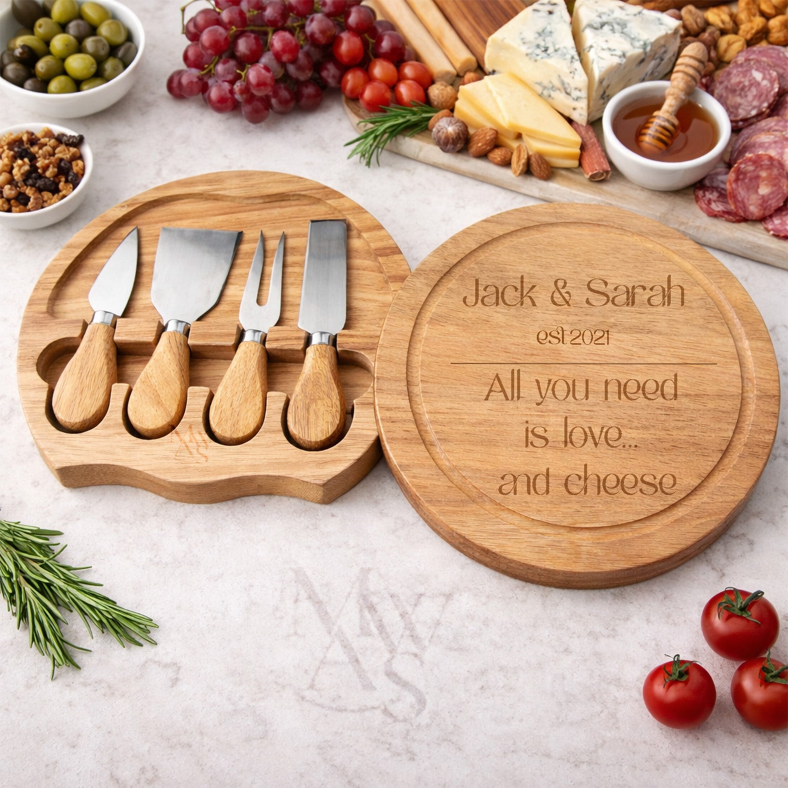 Wooden cheese board set with knives and engraved message on a table with food.