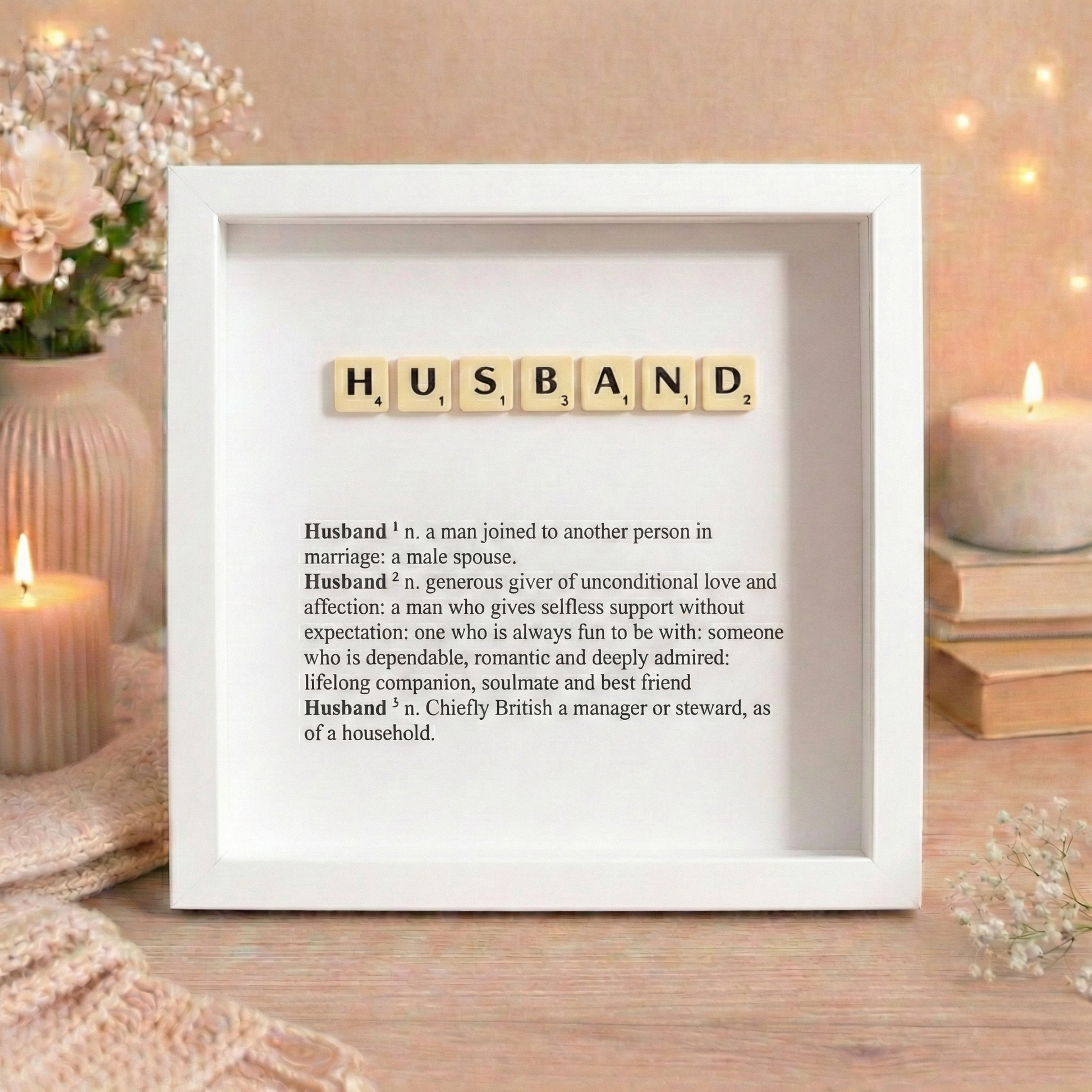 Decorative frame with scrabble tiles spelling 'HUSBAND' and definition on a wooden surface with candles and flowers.