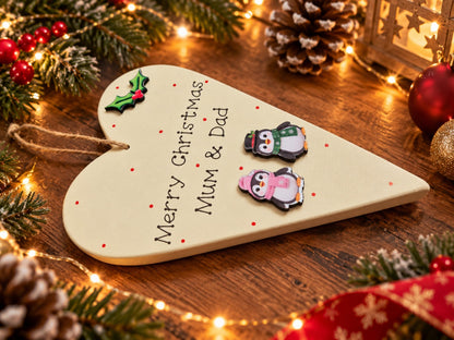 Merry Christmas Mum and Dad wooden heart plaque in penguin design on a festive wooden table with Christmas lights and greenery