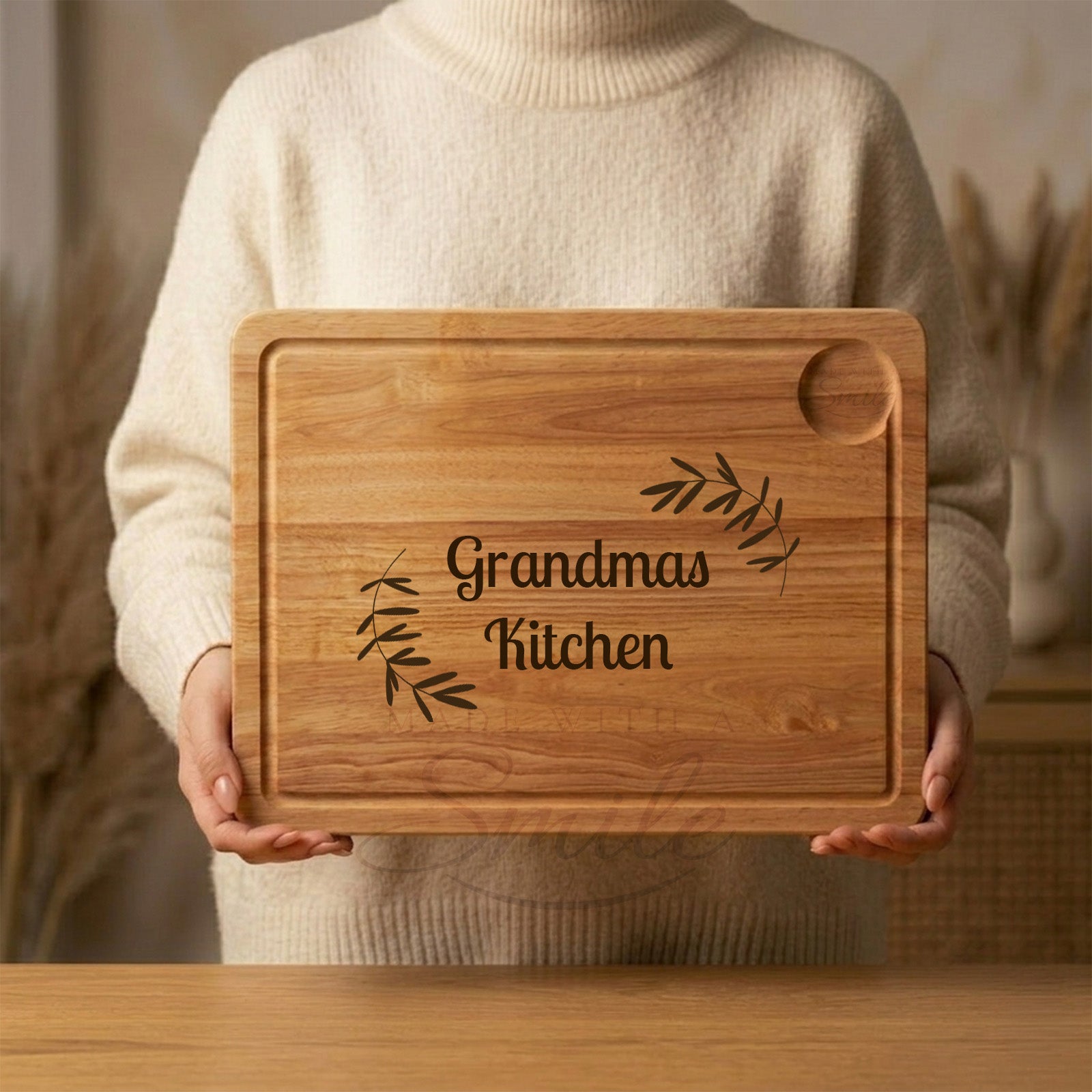 Person holding a wooden cutting board with leaf motif engraving and  'Grandma's Kitchen' engraving.